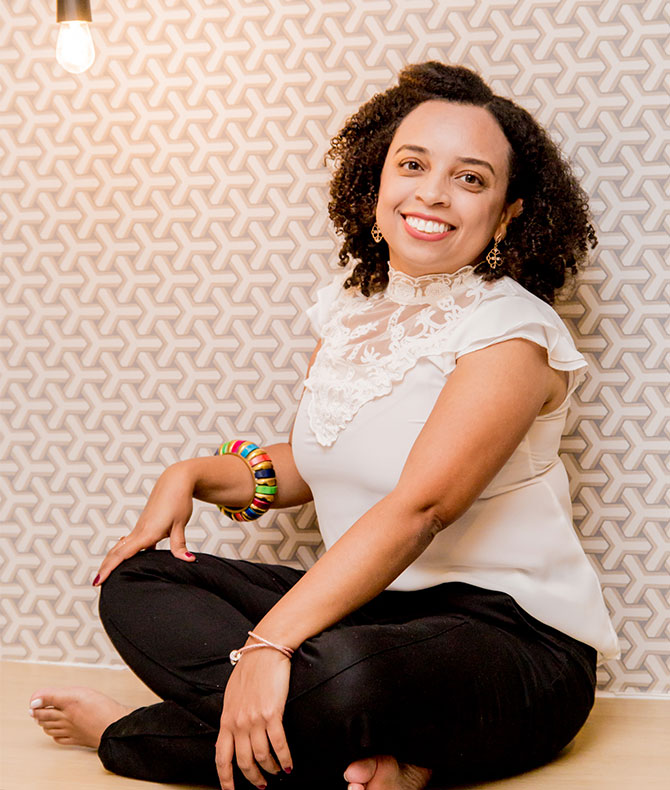 Fotografia de Elaine Rocha sentada no chão de pernas cruzadas, ela veste blusa branca com detalhes e calça preta, um bracelete colorido e está descalça