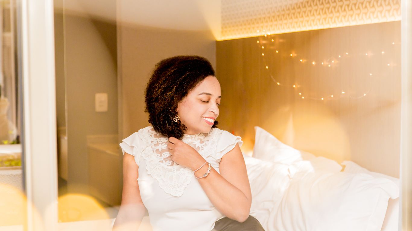 Fotografia de Eliane Rocha sentada em uma cama com travesseiros brancos